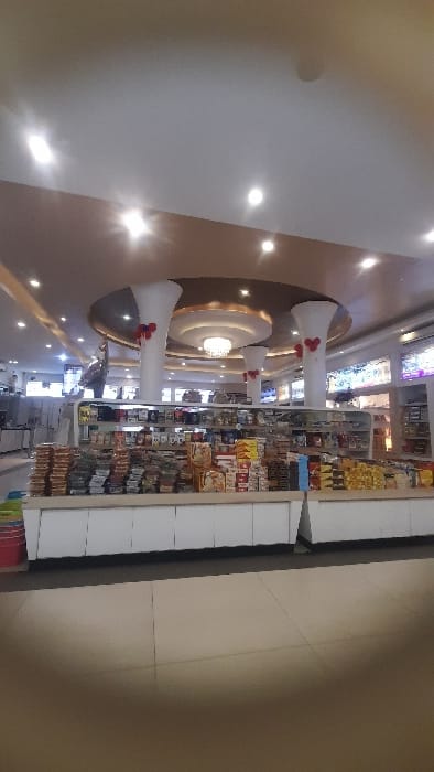 This image depicts the interior of a brightly lit retail establishment located in Glantengan, Indonesia. The scene is devoid of people.
In the midground, a long, multi-level, white display counter dominates the frame, densely packed with various commercially packaged food items, including snacks, biscuits, and confectionery, arranged in organized stacks. To the immediate left of this counter on the floor, a red bucket and a partial blue object are visible.
The background features two prominent, white, fluted columns adorned with red heart-shaped decorations at mid-height. These columns support an ornate, multi-tiered circular ceiling structure with gold accents and a large, multi-bulb chandelier. To the left of the left column, a large, dark-colored, bird-like statue (resembling a turkey or rooster) is positioned on a higher shelf. Further back, additional white display units, refrigeration appliances, and illuminated product shelves are visible on both the left and right sides.
The ceiling is white with numerous recessed downlights, and a central brown/gold decorative bulk-head. The floor is composed of light-toned, square tiles. The entire area is uniformly illuminated by artificial light sources.