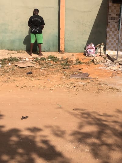 The scene is set outdoors in Egan, Nigeria. A male individual, viewed from the back, stands approximately 0.5 meters from a light green painted wall. He is barefoot, wearing a black t-shirt with a white graphic and bright green shorts. His stance suggests an act of urination or private adjustment. To the right of the individual, the wall transitions to a segment of brown and beige checkered tiles, separated by an orange-brown vertical pillar. Two fabric bags, one pink/white with "3%" visible and another beige/green, rest on the ground near the tiled section. The ground consists of reddish-brown dirt, sparsely covered with dry green vegetation and scattered debris including cardboard pieces, dark plastic, and concrete fragments. Strong daylight is indicated by distinct shadows cast by the individual on the wall and by unseen foliage in the foreground.