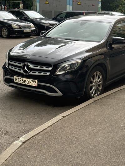 In Zelenograd, Russia, a black Mercedes-Benz CLA-Class is parked on an asphalt road next to a concrete curb. The vehicle's license plate reads "H126BO 799". To the left of the Mercedes, a black Mercedes-Benz E-Class is partially visible. In the background, people are standing near a building with electrical equipment and warning signs. The scene appears to be a typical urban street.