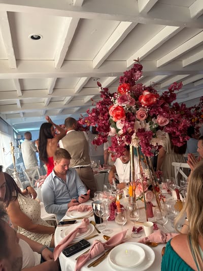 The image depicts an indoor event, likely a wedding reception, held at an unspecified location within the city of Hem, France. A long, white tablecloth covers a dining table adorned with a large, elaborate floral centerpiece featuring red and pink flowers. Place settings include white plates, gold cutlery, and wine glasses. Two lit orange candles are visible on the table.
Several people are seated around the table, engaged in conversation or looking at their phones. A woman in a white polka-dot dress is seated to the left, and a man in a light blue shirt is in the foreground, also looking at his phone. In the background, other guests are visible, with some standing and dancing near a window. The ceiling has exposed white wooden beams, and recessed lighting fixtures are present. The overall atmosphere suggests a festive gathering.