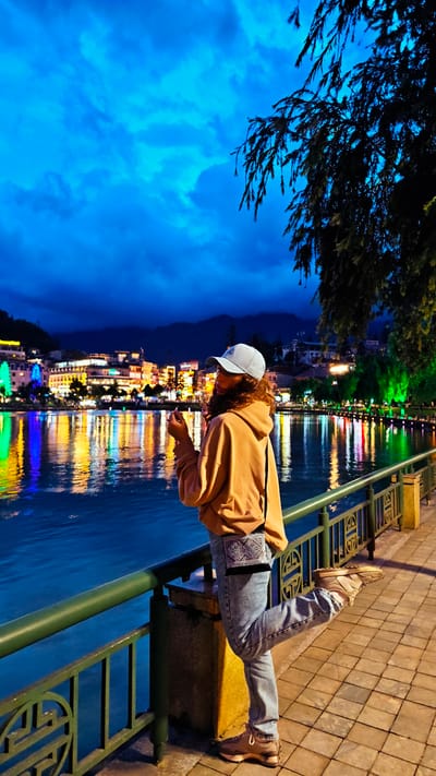 A full-length shot shows a person standing on a sidewalk next to a body of water. The person is female, with curly hair and light brown skin. She is wearing a light blue baseball cap, a tan-colored hoodie, light blue jeans, and tan-colored sneakers. She has a small black and white patterned bag slung across her body. She is leaning against a green metal railing with her right leg raised slightly. She is facing left, looking at the water and the buildings across the way. The water reflects the lights of the buildings. The buildings are brightly lit with various colors. In the background, there are mountains. The sky is a deep blue color, with some clouds. A tree is to the right of the woman. The sidewalk is made of square tiles. The photo was taken in the city of Sa Pa Ward, Vietnam.