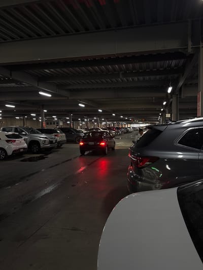 This media file captures a dimly lit, large indoor facility, likely a multi-level parking garage or vehicle storage area. The image is taken from inside a car, looking forward and slightly to the right.
The primary subjects are numerous cars, lined up in rows that extend into the distance. In the immediate foreground, the rear of a dark-colored car with glowing red taillights is visible on the right, positioned next to a white vehicle. Further along the lane, various other cars, including light grey, white, and dark-toned models, are parked or moving slowly, many also displaying their red taillights. The ground beneath the vehicles is a dark, somewhat reflective surface, possibly concrete.
The overhead structure consists of robust industrial-style metal beams and corrugated panels, painted in dark tones. Bright, linear fluorescent lights are affixed to the ceiling, providing the main source of illumination and creating reflections on the car bodies and the floor.
A notable detail is a white sign with a red border visible in the mid-ground on the left, displaying the Spanish word "CIRCULACIÓN" (Circulation), indicating vehicle movement lanes. The overall environment suggests an operational facility, possibly a large port, a vehicle staging area, or a large public parking garage in Cholula, Mexico. The prevalence of artificial lighting indicates either nighttime or a completely enclosed indoor setting. No individuals are clearly visible in the scene.