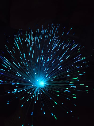 A close-up shot against a black background reveals a cluster of light-emitting fibers. The fibers appear to originate from a central, bright light source, dispersing outwards in a radial pattern. The fibers emit light in shades of blue and green. No people or objects besides the light source are visible. The location is not discernible from the image content.