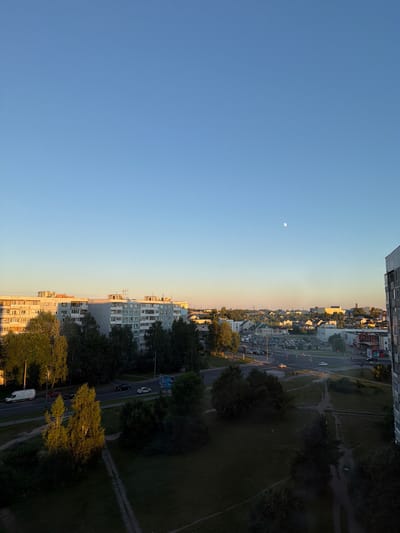 This image shows a panoramic view of a city skyline with a clear blue sky and a faint moon visible. In the foreground, there are residential buildings, some of which are multi-story apartment blocks with many windows. Green trees are scattered throughout the scene, casting shadows on the ground. A road with vehicles, including cars and a white van, runs through the middle ground. In the distance, more buildings and houses are visible, along with what appear to be wind turbines. The overall impression is a suburban or residential area of a city, likely Mahilyow, Belarus, during either sunrise or sunset, given the warm, golden light illuminating the buildings and trees. There are no prominent human figures visible in the image.