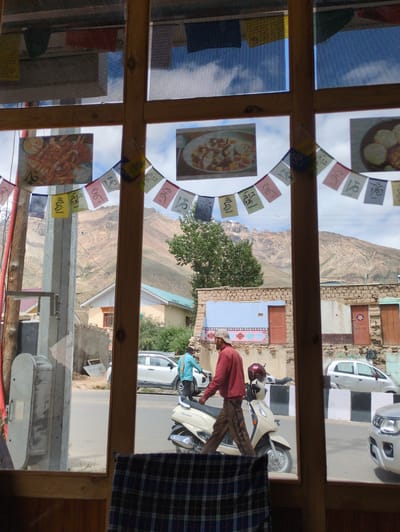A medium shot captures a view through a wooden window frame in Padum, India, overlooking a street scene with traditional buildings and mountains in the background. Prayer flags with Tibetan script are strung across the window.
In the foreground, a portion of a checkered blanket is visible. The scene outside reveals a paved road with a few vehicles, including a white car and a light-colored scooter with a maroon helmet on its handlebars. A person wearing a red jacket and brown pants walks on the road. Another person in a blue shirt is near the white car.
The buildings are constructed with rough stones and some have painted facades. The mountains in the distance are arid and rocky, with sparse vegetation. The sky is cloudy. The overall impression is of a rural or semi-urban setting in a mountainous region of India.