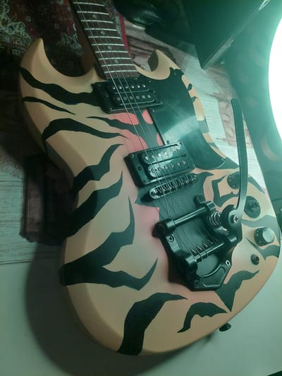 This media file is a close-up, overhead shot of an electric guitar. The guitar has a tiger-stripe pattern with black stripes on a tan background. It features a tremolo system, two black humbucker pickups, and a fretboard with silver frets. The guitar is lying on a surface that appears to be a wooden table or floor, with a rug visible beneath the lower part of the guitar. The lighting suggests it is indoors, possibly under artificial light, as indicated by the bright, somewhat diffused light source at the top of the frame, which also casts a green hue over a portion of the image. There are no people or specific events depicted; the focus is solely on the guitar and its details. The setting is not definitively identifiable beyond being an indoor space, though the provided location context is Markovo, Bulgaria. There is no visible text in the image.