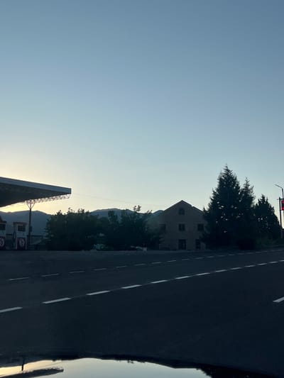 The image depicts a scene at a gas station in Arpi, Armenia, during twilight. A large, angled roof structure, characteristic of a gas station canopy, is visible on the left, supported by metal beams. Two red and white gas pumps are positioned beneath the canopy, with visible branding. Beyond the gas station, a building constructed of light-colored brick with arched windows is present. To the right of the brick building, several tall, dark green coniferous trees are situated. In the background, a mountain range is faintly visible against the clear, pale blue twilight sky. A paved road with white dashed lines runs across the foreground, reflecting a puddle of water in the lower portion of the frame. No people are visible in the image.