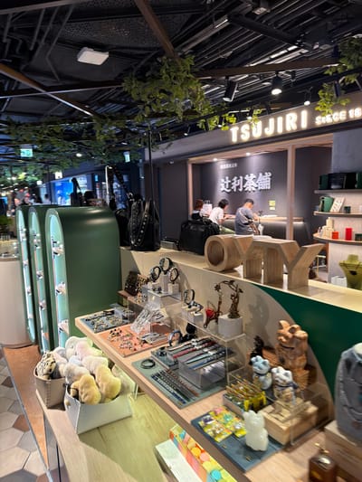 The image depicts the interior of a retail store, likely in Hong Kong, China, as suggested by the presence of Chinese characters on signage. The store appears to be a tea shop, with the brand name "TSUJIRI" prominently displayed in English and Chinese characters. The environment is well-lit, with a modern and slightly rustic aesthetic, featuring wooden elements and hanging greenery.

In the foreground, a display shelf showcases various merchandise. This includes a collection of plush, soft toys in a light-colored woven basket, and another smaller basket with leopard-print fabric. Adjacent to the toys are display cases with an assortment of jewelry and accessories, such as bracelets, necklaces, and watches, arranged on clear acrylic stands. There are also decorative items, including small sculptures.

Further into the store, a counter area is visible with customers and staff interacting. Two individuals are seated at the counter, appearing to be served or engaged in a transaction. One person is wearing a grey t-shirt, and the other a white t-shirt. Another individual is visible in the background, suggesting a service-oriented retail space. Behind the counter, shelves are stocked with products, some of which are in green and white packaging. A large wooden sculpture, possibly spelling out letters, is positioned on a shelf.

The ceiling of the store is a visible industrial-style structure with exposed pipes and wiring, contrasting with the more refined retail display below. Green vines are draped from the ceiling and beams, adding a touch of nature to the indoor environment. Lighting fixtures are strategically placed to illuminate the merchandise and seating areas. The overall impression is that of a contemporary retail establishment offering a blend of traditional and modern elements.