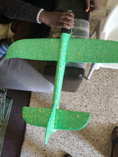 An indoor scene in Nicosia, Northern Cyprus / Cyprus, depicts a person's hands holding a green foam glider toy. The glider, positioned horizontally across the frame, features a green body and wings speckled with small yellow dots, and a black nose cone. A person with dark skin is visible in the upper center, wearing a dark, possibly knitted, long-sleeved garment with a white elastic cuff on the right wrist, gripping the glider's nose cone with their right hand. The glider rests partially on a dark, worn rectangular cushion or armrest. To the right, part of a white plastic high chair or stool with metallic legs is visible. The floor is covered in light-colored, speckled terrazzo or tiled flooring. To the left, a grey upholstered surface, a dark rug, and a green and white checkered textile are present. A dark-colored slipper or sandal is partially visible in the bottom right corner.
