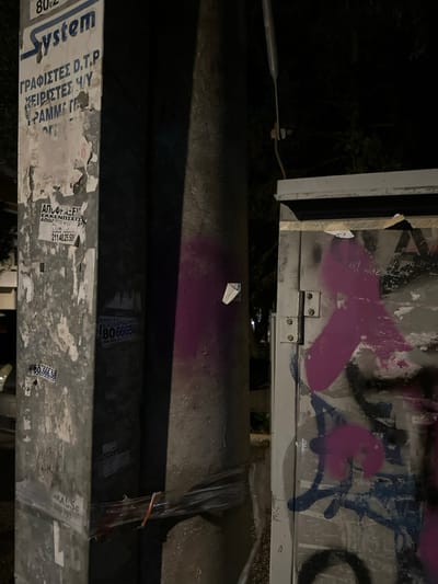 The image captures an upward-looking view of urban infrastructure at night in Marousi, Greece. The scene is dominated by two large, horizontal utility poles or pipes, heavily covered in paper advertisements and graffiti, positioned in the middle and lower parts of the frame. Above these, a metallic structure, possibly a street sign's reverse side or part of a building facade, is visible, featuring bold purple spray paint graffiti and general wear. The overall lighting is dim, indicative of nighttime, with a dark, indistinct background.
There are no people or discernible activities taking place; the image focuses on static, textured urban elements. Notable details include the accumulation of various paper flyers and posters, many partially torn or faded, along the poles. The metallic structure above also shows signs of aging and public markings.
Visible text, primarily in Greek, includes:
- On the lower pole: "System", "ΓΡΑΦΙΣΤΕΣ D.T.P" (Graphic Designers D.T.P), "ΧΕΙΡΙΣΤΕΣ Η/Υ" (Computer Operators), and "ΓΡΑΜΜΑΤΕΙΣ" (Secretaries).
- On a sticker on the mid-level pole: "ΑΠΟΦΡΑΞΕΙΣ" (Drain unblocking services) with phone numbers "211 40.25.509" and "80.66656". Another sticker has "ΑΠΟΦΡΑΞΕΙΣ" repeated.
- A smaller sticker on the upper pole reads "AKA 65".
The purple graffiti on the top metallic structure appears to be abstract. The image conveys a sense of a lived-in urban environment marked by public advertising and street art.