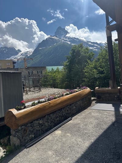 The image, oriented vertically, captures a scenic view of a mountainous landscape with elements of human presence. Dominating the background are majestic, snow-capped mountains under a bright blue sky filled with dramatic white clouds. Lush green trees cover the slopes of the lower mountains, contrasting with the rugged, rocky terrain above. In the mid-ground, several buildings are visible, including one with a green roof and another appearing to be under construction with scaffolding. A yellow crane stands near the buildings, suggesting ongoing development or renovation.

In the foreground, a wooden planter box filled with rocks and small, colorful flowers adds a touch of domesticity to the wild landscape. The planter is constructed from a large, hollowed-out log. Adjacent to this planter is a paved area, likely a walkway or patio, with a shadow cast across it by an unseen structure, indicating it is daytime, likely late morning or early afternoon. The overall mood is serene and picturesque, highlighting the beauty of nature alongside signs of human activity. The location is identified as Dombay, Russia. No visible text is present in the image.