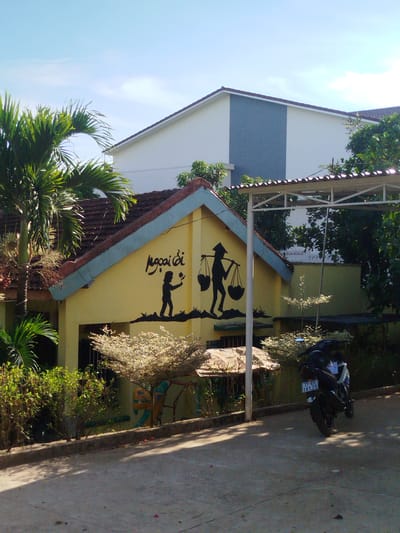 The scene is an exterior view in An Phu Ward, Vietnam, featuring a yellow one-story building with a red-tiled, gable roof. On the building's facade, there are black silhouette images: a child holding a flower, and a figure with a conical hat carrying baskets on a yoke. Next to the building is a taller, two-story building painted white with a gray vertical stripe. A palm tree stands to the left, with other trees and shrubbery in front and along the side of the building. A motorcycle is parked under a metal canopy structure to the right of the yellow building, showing its license plate, which reads "77-E1 191.25". The scene is bathed in sunlight, casting shadows on the concrete ground.