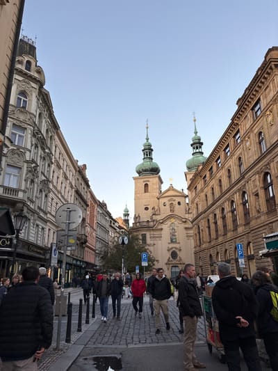 The image depicts a bustling cobblestone street in Capital City of Prague, Czechia, specifically Mostecká Street looking towards the Church of Saint Nicholas in Malostranské náměstí. The Church of Saint Nicholas is centered in the background, a prominent Baroque structure with two tall, tan bell towers topped by green onion domes, spires, and crosses. Its central facade is highly ornate, featuring statues and a large window. Above, the sky is clear and light blue.

On the left, a continuous line of multi-story historical buildings with varied, ornate facades in shades of cream, grey, and pinkish-orange extends into the distance, with retail shops on their ground floors. A distinctive corner building with a small turret is visible at the far left. On the right, a similar multi-story building with a light brown stone facade and arched windows, some adorned with gold medallions, also hosts ground-level businesses.

The cobblestone street is occupied by approximately 20-30 pedestrians, mostly adults, walking in both directions. They are dressed in casual fall/winter attire, including jackets and long pants. In the foreground, a person in a black puffer jacket stands with their back to the camera. Further into the scene, a man in a red jacket walks towards the viewer, while a person stands next to a multi-tiered pushcart on the right side of the street. Black bollards line the pedestrian areas. Various street signs, including blue 'P' parking signs and circular informational signs, are visible on poles, along with a double-faced street clock positioned mid-ground to the left of the church. A small puddle is visible on the cobblestones in the left foreground.