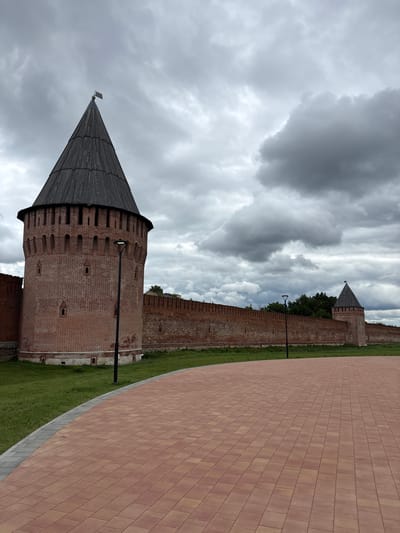 The image shows a view of an old fortress with brick towers and walls. The fortress is surrounded by green grass, and a paved walkway runs along the side. The sky is cloudy and overcast, suggesting a cool or rainy day. In the foreground, a large conical tower with a wooden roof dominates the left side of the frame. Further back, another tower and part of the fortress wall are visible, with trees growing behind them. The walkway is made of reddish-brown paving stones. There are no people visible in the image. The scene appears to be a historical site or park, possibly in Lubnya, Russia, given the architectural style of the fortress. The overall mood is somber due to the cloudy sky and the imposing nature of the fortress.