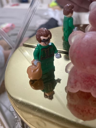 The image shows a LEGO minifigure, identifiable as Dr. Octopus, lying sideways on a highly reflective, golden surface. The minifigure is primarily green with gold detailing on its torso, brown hair, and black goggles over its eyes. Brown, satchel-like appendages, representing the character's mechanical arms, are visible, with one having a grey tip. A clear reflection of the minifigure is cast onto the gold surface beneath it.

Above and partially obscuring the minifigure are several translucent, pink and reddish spherical objects, resembling candies, gumballs, or decorative jelly-like spheres. Some of these spheres have small red specks. These objects are also subtly reflected on the gold surface. In the foreground, a clear plastic or glass barrier runs diagonally, adding another reflective element to the scene. The background is out of focus, revealing indistinct, light-colored shapes typical of an indoor setting.

The scene depicts a static display, possibly part of a decorated cake or dessert arrangement. There is no visible text in the image. The setting for this display is in Ihtiman, Bulgaria.