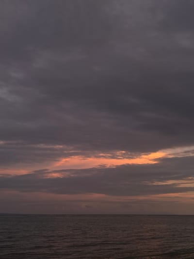 The image depicts a seascape taken at Sainte-Anne, France, featuring the ocean on the right side and a cloudy sky on the left. The clouds are a mix of dark grey and light orange, suggesting either a sunrise or sunset. The sea appears relatively calm with dark, reflective water. There are no people or objects visible on the water or beach.