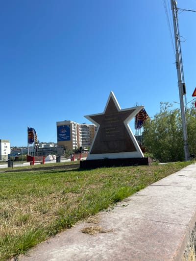The image shows a monument in the shape of a star in Yakutsk, Russia. The monument has inscriptions in Russian. In the background, there are several buildings, including a tall apartment building with a large banner. Two flags with orange and black stripes are displayed on flagpoles. A street light pole with wires is visible on the right side of the image. The ground is covered with grass and concrete. The sky is clear and blue.