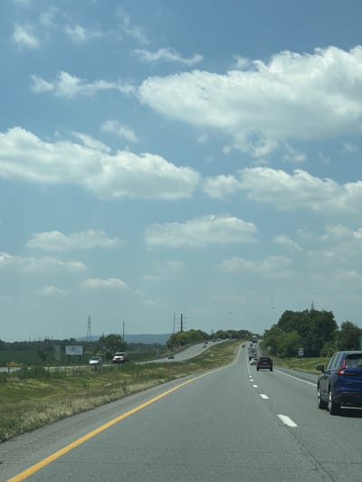 The image captures a highway scene in West Hempfield Township, United States of America, under a bright, cloudy sky. The perspective is from a vehicle traveling on the road, looking forward. The highway has multiple lanes and a yellow line dividing the lanes on the left side. Various vehicles are visible on the road, including a blue SUV in the foreground on the right, a red SUV further ahead, and other cars and trucks dispersed along the highway. The surrounding environment consists of green grassy areas with some vegetation and trees on the sides of the road. In the distance, power lines and transmission towers are visible, along with rolling hills. A billboard is also present on the left side of the highway. The overall scene depicts a typical daytime highway commute.
