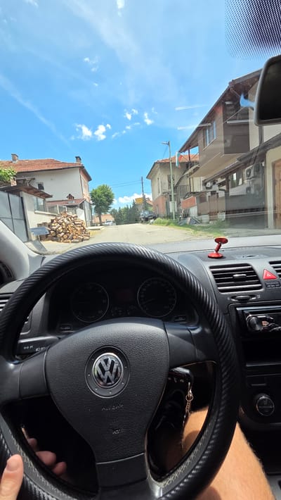 A first-person perspective from inside a car shows a road lined with houses and trees under a bright blue sky with scattered white clouds. The steering wheel, with a Volkswagen logo, is visible in the foreground. To the right, a dashboard with an air vent and a red suction-cup accessory is present. The car is parked on a street in the city of Bania, Bulgaria, as indicated by the Bulgarian flag hanging from one of the houses and the architecture typical of the region. A pile of chopped firewood is stacked on the left side of the road.