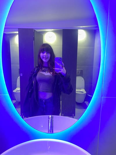 A young woman takes a mirror selfie in a public restroom. She wears a black jacket, jeans, and a light top. Her long nails are visible as she holds a purple phone. Reflected in the illuminated oval mirror is a toilet, a row of lights, gray tile, and sinks. Blue light dominates the scene, emanating from the mirror's frame.