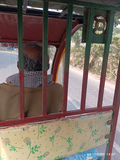 The image depicts an interior view from a moving three-wheeled vehicle, likely an auto-rickshaw, in Kaliganj, Bangladesh. The foreground is framed by the vehicle's structural elements, consisting of green vertical and red horizontal metal bars. A worn cream-colored seat cushion with a faded green leaf and rose pattern is visible at the bottom, partially obscuring a blue floral cushion. To the upper right, a rusted green metal housing, possibly for a fan or light, is attached to the frame. A red canopy or roof component is visible at the top.
An adult individual with short, disheveled grey hair is seated in the vehicle, viewed from behind. The person is wearing a light brown garment and a grey and white checkered scarf.
Through the vehicle's opening, a paved road is visible, suggesting movement. The road is bordered on the right by a dirt shoulder and dense green tropical foliage, including trees and undergrowth. The scene is illuminated by natural daylight. A white sticker with red Bengali script is affixed to the vehicle's interior on the left side. A digital timestamp "04/12/2025 13:52" is present in the lower right corner of the image.