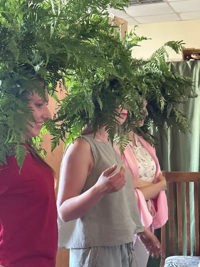 The image shows two women interacting amongst a large arrangement of ferns. The woman in the foreground, wearing a red shirt, is looking upwards and smiling. The second woman, wearing a pale green top and a pink shawl over a floral dress, is lying down, with her head propped on a pillow. Her eyes are closed, and she appears to be resting. The ferns are dense and green, framing the faces of both women. The scene is set indoors, with a tiled wall visible in the background, suggesting a home or a studio environment. A wooden chair with slatted back is also partially visible. The lighting suggests daytime, with natural light filtering in. There is no visible text in the image. The overall mood is peaceful and intimate, with the lush greenery creating a serene atmosphere. The people are positioned such that the ferns create a natural canopy above and around them, giving the impression of a secluded, natural haven within an indoor space.