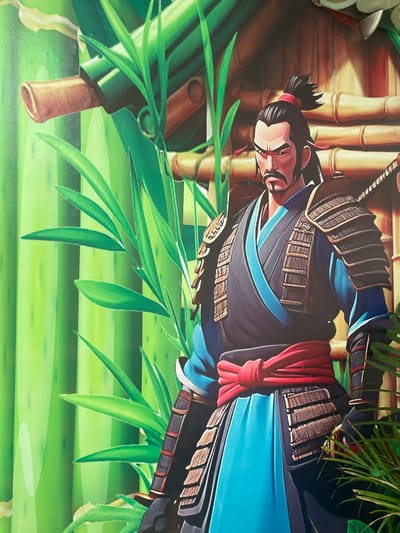 The image shows a painting of a samurai warrior set against a backdrop of bamboo stalks and leaves. The samurai is depicted wearing traditional armor and a topknot hairstyle. The colors are vibrant, with shades of green, brown, blue, and red dominating the scene.

I do not have information about the location of the image.