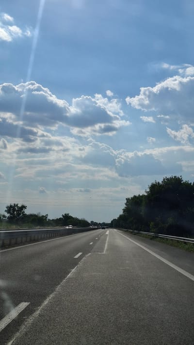 The image captures a perspective from inside a vehicle traveling on a highway. The road stretches forward, divided by white lines, with trees and vegetation lining both sides. In the distance, other vehicles can be seen on the road. Above, a bright blue sky is filled with dramatic, white cumulus clouds, with a sunbeam piercing through them from the upper left. The scene appears to be set in an outdoor environment, possibly a rural or suburban area, given the presence of trees and open road. The specific location in Войводино, Bulgaria cannot be determined from the visual information alone.