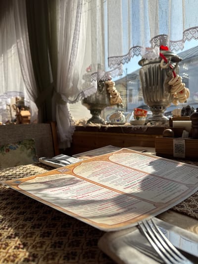 Here is a description of the image:
The image depicts a restaurant or cafe setting. A menu is prominently displayed, detailing various dishes, including soups, dumplings, and main courses, with prices listed in Russian currency. In the background, there's a samovar, a traditional Russian tea urn, indicating the establishment's cultural or historical theme. Light streams in through a window covered with a lace-trimmed curtain. The table is covered with a patterned cloth. The image evokes a sense of traditional Russian dining. The mention of "Ростовская" in the menu suggests the restaurant or establishment might have a specific regional culinary focus.