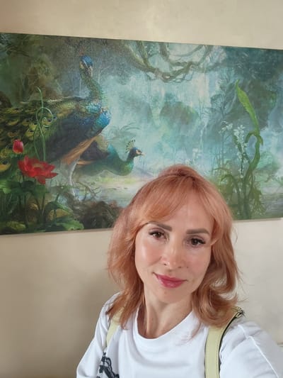 A selfie shows a woman with reddish-blonde hair and makeup posing in front of a painting. The painting depicts two peacocks in a lush, misty forest setting with red flowers and white blossoms. The woman is wearing a white t-shirt with a black lace design on the bottom and yellow straps over her shoulders. She has a slight smile and is looking towards the camera. The background behind the woman is a plain, light-colored wall. There are no visible text elements in the image. The scene appears to be indoors, likely in a home or gallery, given the artwork on the wall. The lighting is soft, suggesting it could be daytime.
