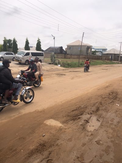 In Hanwa, Nigeria, the image captures a street scene with a dirt road. Two motorcycles are visible; one is parked with multiple riders, while the other is further down the road. Adjacent to the road is a row of parked vehicles, including a van and cars. Buildings are visible in the background, interspersed with trees and utility poles. The sky is overcast.