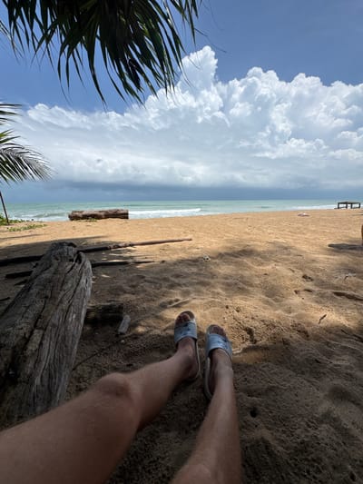 The image depicts a beach scene in Ban Khuk Khak, Thailand. In the foreground, a person's legs are visible, wearing light blue slides, resting on the sandy beach. To the left, a large piece of weathered driftwood is prominent, with more scattered pieces of wood on the sand. Palm fronds hang over the top left of the frame, casting shadows on the sand. The beach extends towards the ocean, which has moderate waves under a partly cloudy sky. In the distance, a wooden structure resembling a pier or seating area is visible on the right side of the beach. The overall environment is a tropical coastal setting.