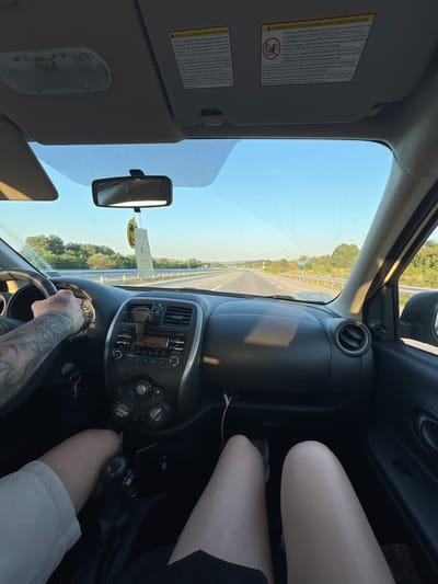 This is a first-person perspective video captured from inside a car traveling on a highway. The subjects are people, visible from their legs and arms, and various car interior objects like the steering wheel, dashboard, radio, and rearview mirror. The scene is set on a sunny day, with a clear blue sky and green trees lining the road. The activity is driving on a highway in Bulgaria, as suggested by the location context and the visible text on the radio display showing "BG RADIO." The time of day appears to be daytime, likely late morning or afternoon, given the bright sunlight and shadows. The overall mood seems relaxed, suggesting a road trip or a casual drive. The dashboard has several controls and displays, including air vents and buttons for the car's audio system. The sun visor is visible in the top left, with warning labels containing text. A hanging air freshener is also visible.