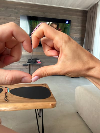 Two hands are forming a heart shape in the foreground. The hands are close together, with the thumbs and index fingers touching to create the top curves of the heart, and the other fingers extending downwards to form the point. The skin tone is light, and the fingernails on the right hand are painted with a light pink polish. In the background, a television screen is visible, showing a person's face and part of a pink garment. To the left of the television, a wooden wall paneling is present. A white curtain hangs on the right side of the image. Below the hands, a wooden table with black hairpin legs is visible. On the table, there are two black oval placemats and a pair of Nintendo Switch controllers, one orange and one blue, attached by a black strap. The floor is a light, neutral color. A portion of a light-colored sofa is visible on the far right of the frame. The image appears to have been taken indoors, possibly in a living room. The location is not explicitly stated in the image, therefore no information about Zarozhdeniye, Russia can be provided.