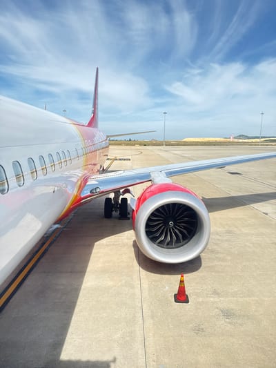 An airplane engine, painted red and silver, is in the foreground on the tarmac at Cam Ranh International Airport in Cam Ranh, Vietnam. The airplane's fuselage, white with red and yellow accents, extends to the left. The wing, also silver, stretches out to the right. A small orange and yellow traffic cone sits on the concrete ground in front of the engine. In the background, a large pile of sand and construction equipment are visible, along with distant hills under a bright blue sky with wispy clouds. The scene is brightly lit by the sun, casting long shadows.
