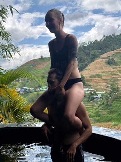 A female stands on the shoulders of a male within an outdoor hot tub, both subjects appearing wet, against a mountainous landscape in Mù Cang Chải, Vietnam. The female, positioned upright and facing left with a smile, wears a black sports bra displaying a "NIKE" logo and black bikini bottoms; a tattoo is visible on her left arm. The male, partially submerged in the water, supports the female on his shoulders, his head turned right with a pensive expression. He is wearing dark swim shorts. Green foliage, including palm fronds and bamboo, partially frames the left side of the scene. The background features extensive agricultural rice terraces covering steep hillsides, interspersed with small residential structures, under a partly cloudy sky with visible blue patches.