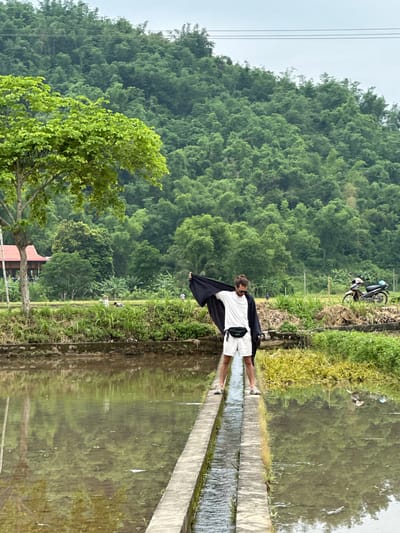 A young man with a beard and sunglasses stands on a narrow concrete path between two flooded rice paddies in Mai Chau, Vietnam. He wears a white t-shirt, white shorts, and a black fanny pack. He is holding a dark blue or black garment draped over his shoulders and his arms. In the background, lush green trees cover a hillside. A motorbike is parked on the right side of the scene. The sky is overcast. The water in the paddies reflects the surrounding greenery.