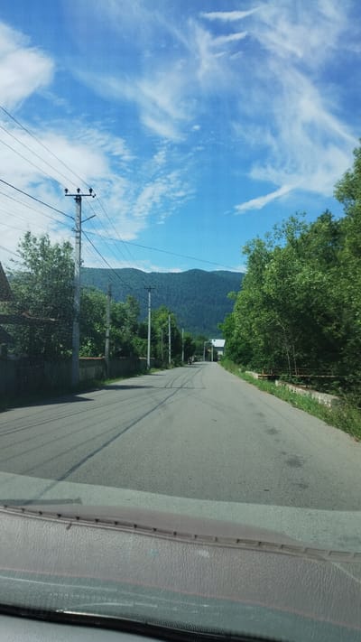 The image shows a view from inside a vehicle looking out at a paved road in Mizhhiria, Ukraine. The road curves to the right and leads towards a densely forested mountain range in the distance. Overhead, a bright blue sky is dotted with wispy white clouds. On either side of the road, there are trees and houses, and power lines run along poles. The foreground of the image shows the dashboard of the vehicle.