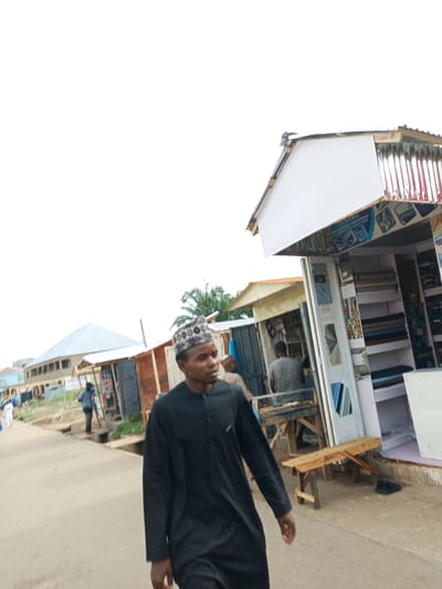 In Arawa, Nigeria, a busy marketplace scene unfolds. A man in a black traditional outfit and a patterned cap walks down a paved street, his gaze directed forward. Behind him, a row of open-air shops line the street, displaying textiles and various goods. In one shop, rolls of fabric are neatly arranged on shelves. Another man, wearing a grey t-shirt and dark pants, is visible inside a shop, possibly attending to customers. The environment is a typical urban setting with buildings and a clear, overcast sky. The street appears to be a thoroughfare for both pedestrians and possibly vehicles, though none are prominently featured in this particular shot.