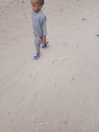 A young boy is walking on a sandy surface. He wears a gray outfit and blue sandals with floral decorations. The ground is covered with sparse pieces of debris, possibly dried twigs or leaves. He is walking to the left of the frame, looking in the same direction. The background appears to be an open, dry area.