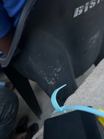 The image depicts a close-up, low-angle view of a large black plastic object, possibly a piece of furniture or equipment, with white lettering that appears to read "BUS." The object has visible scuff marks and scratches on its surface. In the foreground, a bright blue plastic item, possibly a hanger or a similar accessory, is positioned horizontally across the frame, resting on a rough, speckled surface, likely concrete or stone. To the left and bottom of the frame, parts of a person's legs and feet are visible, wearing dark-colored pants and shoes. The overall environment appears to be outdoors, with subdued lighting. The location in Mayo Dasa, Nigeria, is not discernable from the visual information provided in the image.