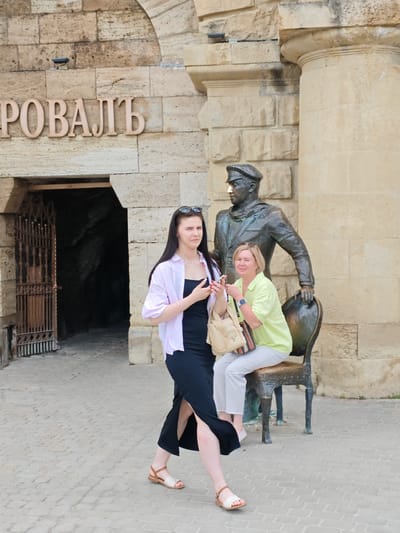 Two women are posing for a photo in front of a stone building and a statue. The woman on the left is wearing a black dress with a slit up the side, a light pink open shirt, and sandals. She is holding a phone in her hand. The woman on the right is sitting on a statue of a man in a uniform who is seated on a chair. She is wearing a light green shirt and white pants and is also holding a phone. She is smiling. The statue is made of bronze and depicts a man wearing a cap and uniform, with a scarf around his neck. The building behind them is made of large stone blocks, and above an entrance, there is text that reads "ПОРОВАЛЬ". The entrance appears to lead into a dark cave-like area with a wrought iron gate. The ground is paved with cobblestones. The scene appears to be outdoors on a bright, likely daytime, setting. The overall mood seems casual and tourist-like.