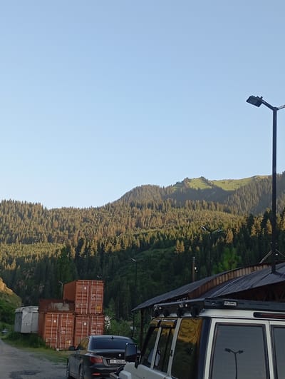 A landscape view captures a vast, treed mountain range under a clear, pale blue sky. The mountains, densely covered with dark green coniferous trees, rise from the valley floor. The sun casts long shadows, indicating either early morning or late afternoon. In the foreground, a dark sedan is parked on a dirt road. Behind the car, a stack of orange shipping containers is visible, along with a silver trailer. To the right, the roof of a building with a dark, corrugated metal roof and wooden accents is partially visible. A modern street lamp stands tall on the right side of the frame, silhouetted against the sky. Another street lamp is visible reflected in the window of a white vehicle, which is parked in the foreground and partially cropped out of the image. The scene appears to be in a rural or semi-industrial area situated at the base of a mountainous region. The license plate on the dark sedan reads "01 060 BM".