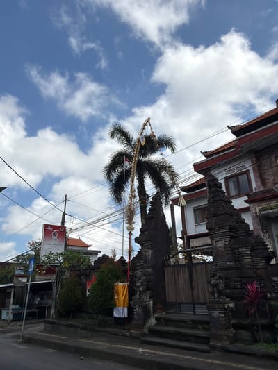 The image depicts a street scene in Tampaksiring, Indonesia. In the foreground, a paved street curves to the left, with a concrete curb running along its edge. Behind the curb is a sidewalk or small area of vegetation. To the right, a traditional Balinese stone gate with carved details and figures guards an entrance to a building. The gate is flanked by stone pillars. A wooden gate with a decorative metal top is visible behind the stone structure. Steps lead up to this gate.
A tall palm tree with a slender trunk stands behind the gate, adorned with festive decorations including a red and white flag and streamers. Behind the palm tree and gate, a multi-story building with a red tiled roof and white walls is visible. It features dark framed windows and intricate roof eaves.
To the left of the gate and palm tree, a street sign is mounted on a pole. The sign has red and white elements and appears to contain text in Indonesian. Electrical wires are strung across the scene, supported by poles, indicating an urban environment. Further to the left, a small stall or shop with a dark roof is present. In the middle ground, there are trees and shrubs.
The sky above is a bright blue, dotted with white, fluffy clouds. The overall impression is of a sunny day in a Balinese town.