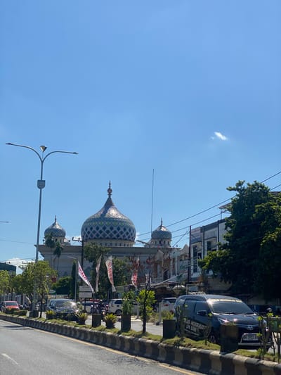 A street view on a clear day shows a mosque with multiple domes in the background. The largest dome is prominent, decorated with a diamond pattern in blue and green. Several cars are parked or driving on the street. A black minivan in the foreground has text printed on its windows. On the left side of the road, a street lamp with multiple arms stands tall. The street is lined with a curb and decorative plants. Buildings, trees, and power lines are visible on the right side of the street. The sky is a bright, clear blue with a small cloud. This scene appears to be in a city in Jawa, Indonesia.