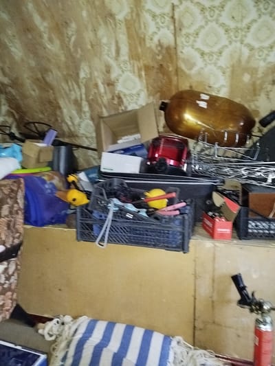 The image depicts a cluttered indoor environment, possibly an attic or storage room, with various objects scattered around. The walls are covered with patterned wallpaper, appearing somewhat aged and discolored.
Prominently featured is a large, oval-shaped, golden-brown object, possibly a gas tank or a decorative item, resting on a metal rack. To its left, a red appliance, likely a toaster or food processor, is visible. In the foreground, a blue plastic crate is filled with miscellaneous items, including what appear to be tools or small household objects, such as a yellow object with a red handle and pink items.
Other items in the scene include cardboard boxes, plastic bags, a blue fabric, and a striped blue and white textile item lying on the floor, partially unrolled. A fire extinguisher with a red cylinder and a black nozzle is positioned in the lower right corner. The overall impression is one of disarray and storage of various materials.
There is no clear indication of the specific location within Дзешчанскі сельскі Савет, Belarus, as no identifying landmarks or signs are visible. The scene is static, with no people or discernible actions taking place.