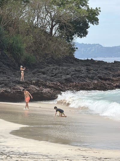 In a tropical beach scene at Manggis, Indonesia, three individuals are enjoying a day by the ocean. The sun is high, indicating it's likely midday, and the weather appears to be clear. The foreground is dominated by a sandy beach with gentle waves lapping at the shore. To the left, a dark, rocky outcropping covered in lush green foliage rises up, providing a stark contrast to the soft sand and turquoise water.
Two people are visible on the sandy part of the beach. One, wearing orange shorts, stands with his back to the camera, arms outstretched as if posing or enjoying the moment. Further down the beach, closer to the water, a second person is seen from behind, wearing a dark shirt and shorts, appearing to be interacting with the shallow water. Near the rocky area, a third person is standing, holding a phone, possibly taking a photo or video.
The overall mood seems relaxed and carefree, with people enjoying the natural beauty of the beach. The clear sky and bright sunlight suggest a warm and pleasant day for outdoor activities. The presence of the rocky cliff and the dense vegetation hints at a wilder, more natural landscape surrounding the beach. No visible text is present in the image.