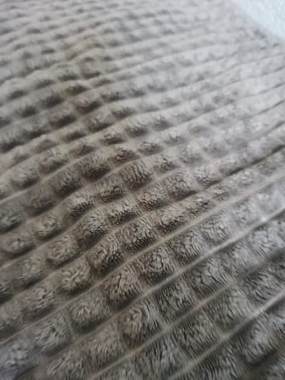 The image displays a close-up, angled perspective of a textured textile surface, observed in an unspecified indoor location in Pokrovnik, Bulgaria. The fabric exhibits a uniform grayish-brown coloration with a plush or fleece-like texture. A repeating pattern is evident across the surface, composed of raised, approximately square or rectangular blocks arranged in diagonal rows. These raised segments are separated by recessed, narrower linear channels. The texture within the raised blocks appears uneven and fibrous. The depth of field is shallow, rendering the foreground in sharper focus and areas towards the upper portion of the frame slightly blurred. No individuals, animate subjects, or additional distinct objects are present within the frame, nor are any actions or interactions depicted.