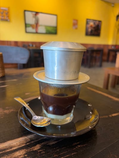 A close-up shot shows a Vietnamese phin filter coffee brewing into a clear glass cup on a dark wooden table. The phin filter, made of silver-toned metal, sits atop the glass, with coffee grounds visible inside. The brewed coffee appears dark, layered with a lighter liquid, likely condensed milk, at the bottom of the glass. A small, shiny silver spoon rests on a dark saucer beneath the cup.

In the background, a bright yellow wall is adorned with two framed pictures. The first picture, to the left, is a blurred image of what appears to be an abstract artwork. Below it, a smaller, orange-toned rectangular sign is visible. To the right of the coffee, the wooden table has a textured, worn appearance. Further in the background, some furniture, possibly chairs, and wooden elements of the establishment's interior are dimly lit. The overall lighting suggests an indoor setting, possibly a cafe or restaurant. The image appears to be taken during the day, given the ambient light. The scene evokes a sense of a quiet moment, possibly the enjoyment of a freshly prepared beverage. No visible text is legible in the image.
