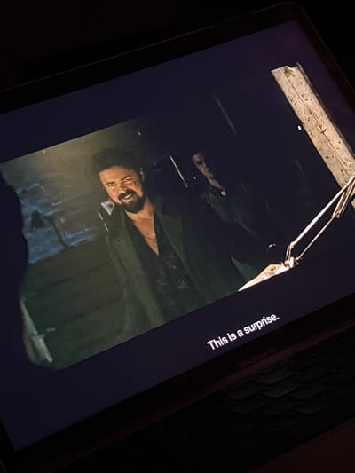 A slightly angled shot shows a tablet screen displaying a scene from a film or show. A man with a beard and a pained expression is visible. Text on the right reads "This is a surprise." Parts of a keyboard and some other ambient light are visible, indicating the shot was likely taken in a dimly lit indoor setting. The image does not contain any information to deduce the location.